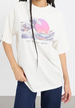 Osoba ubrana w biały T-shirt z nadrukiem fioletowo-różowej fali i słońca oznaczonym "Wielka Fala", w połączeniu z ciemnoniebieskimi dżinsami.