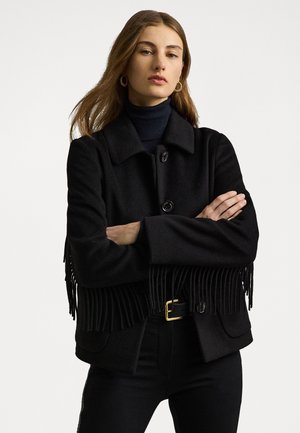 Black wool coat with fringed hem, collared design, front button closure, and two front pockets. Paired with fitted black trousers and a turtleneck.