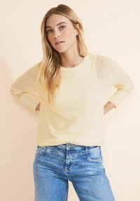 Hellgelbes Strickpullover mit langen Ärmeln und strukturiertem Muster, kombiniert mit blauen Jeans. Weicher Stoff und runder Halsausschnitt sind sichtbar.