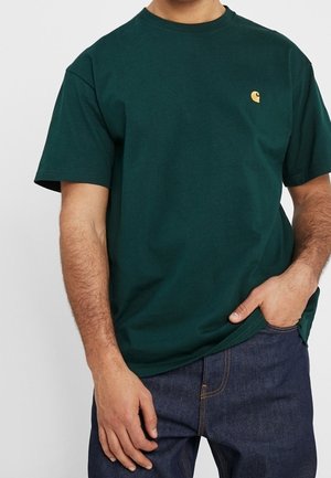 Homme portant un t-shirt vert foncé à manches courtes avec un petit logo jaune sur la poitrine et un jean bleu foncé, une main dans la poche.