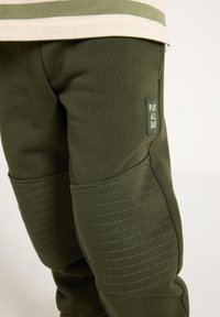 Ελαιούχα πράσινα παντελόνια φόρμας με ραμμένες λεπτομέρειες και υφασμένο πάνελ στο γόνατο. Διαθέτουν ορθογώνιο έμπλαστρο με το κείμενο "ΠΑΙΞΕ ΟΛΗ ΜΕΡΑ" σε λευκό χρώμα.