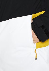 Veste colorée avec des sections noires, blanches et jaunes. Comprend une poche zippée et un cordon de serrage réglable à l'ourlet.