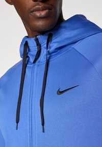 Mėlyna sportinė striukė su minkšta medžiaga, su juodu užtrauktuku ir raišteliais, o krūtinėje yra mažas juodas Nike logotipas.