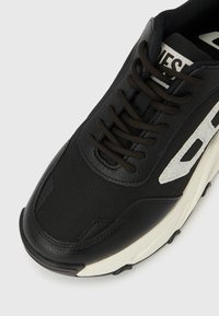 Μαύρο αθλητικό παπούτσι με λευκή σόλα, μαύρα κορδόνια και λευκή γραφική λεπτομέρεια στο πλάι, εμφανιζόμενο σε κοντινή λήψη από μπροστινή γωνία.