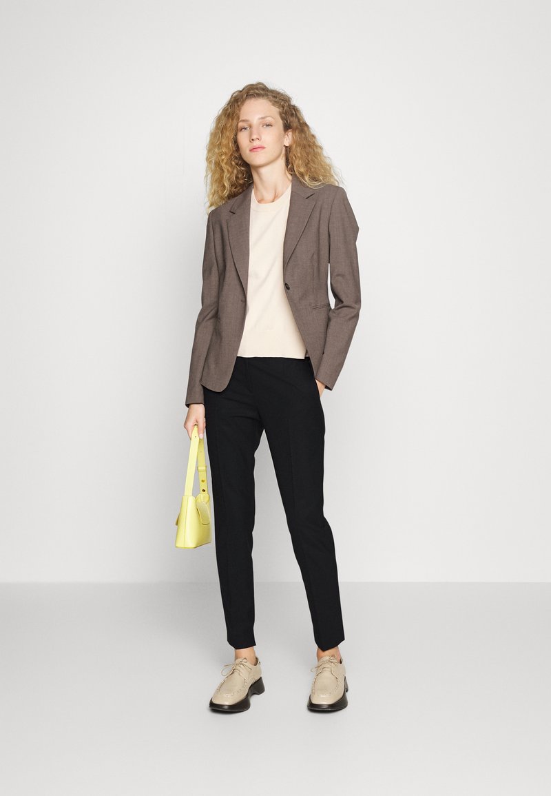 Brauner Blazer über einem beigen Oberteil, schwarze enganliegende Hose und beiges Schuhwerk. Hält eine kleine gelbe Handtasche. Schlichter Hintergrund.