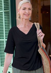 Schwarze, drapierte Bluse mit kurzen Ärmeln und V-Ausschnitt. Kombiniert mit hellen Hosen und einer gewebten Tasche mit mehrfarbigen Streifen.