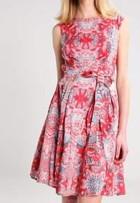 Rotes Kleid mit enganliegendem Oberteil und weit schwingendem Rock, das komplizierte florale und geometrische Muster in Blau- und Rosatönen zeigt. Glatte Textur.