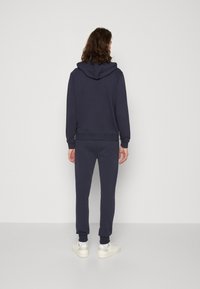 Tmavě modrá mikina s kapucí, kterou lze upravit, doplněná o odpovídající tepláky. Měkká látka s žebrovanými manžetami a pasem, doplněná bílými teniskami.