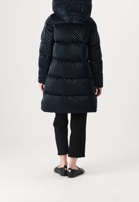 Granatowa pikowana kurtka w kratę z kapturem, z wzorem pikowania, zestawiona z czarnymi krótkimi spodniami i czarnymi płaskimi butami.