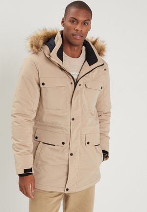 Beige Parka mit abnehmbarem Kunstfell-Kapuze, ausgestattet mit vier Fronttaschen, schwarzen Akzenten und einem Reißverschluss. Weiche Stofftextur.