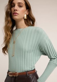 Světlé zelený žebrovaný pletený svetr s dlouhými rukávy, doplněný hnědým páskem, zlatými šperky s akcentem a hnědými koženými kalhotami.