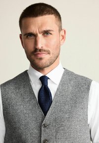 Gray wool vest with a notch collar, featuring three buttons and a textured navy tie, worn over a white dress shirt. Solid background.