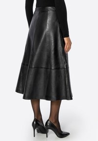 Jupe midi en cuir noir avec une silhouette évasée, présentant des coutures pour la structure et une texture lisse et brillante. Portée avec des talons noirs.