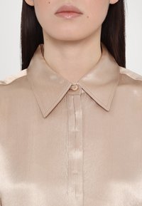Gros plan d'une personne portant une chemise boutonnée en soie beige avec un col pointu et des lèvres brillantes visibles.