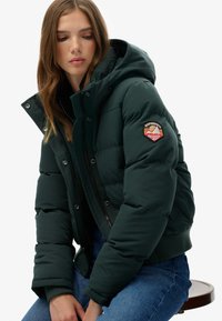 Mørkegrøn puffjakke med hætte, der har knap- og lynlåslukning, ribbede manchetter og et cirkulært broderet mærke på ærmet.