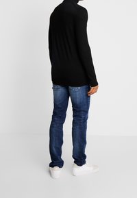 Maglione nero a collo alto con maniche lunghe, abbinato a jeans in denim blu e sneaker bianche, visto da dietro. Design pulito e minimale.