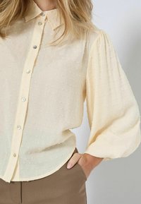 Hellgelbe Bluse mit strukturiertem Punktemuster, Knopfleiste und langen, gepufften Ärmeln. Hergestellt aus einem weichen, durchsichtigen Stoff.