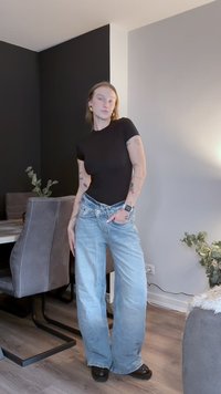 Μαύρο στενό μπλουζάκι, ανοιχτό μπλε παντελόνι με φαρδιά πόδια και μαύρα παπούτσια. Εσωτερικός χώρος με γκρι και μαύρους τοίχους, ξύλινο πάτωμα και φυτά.