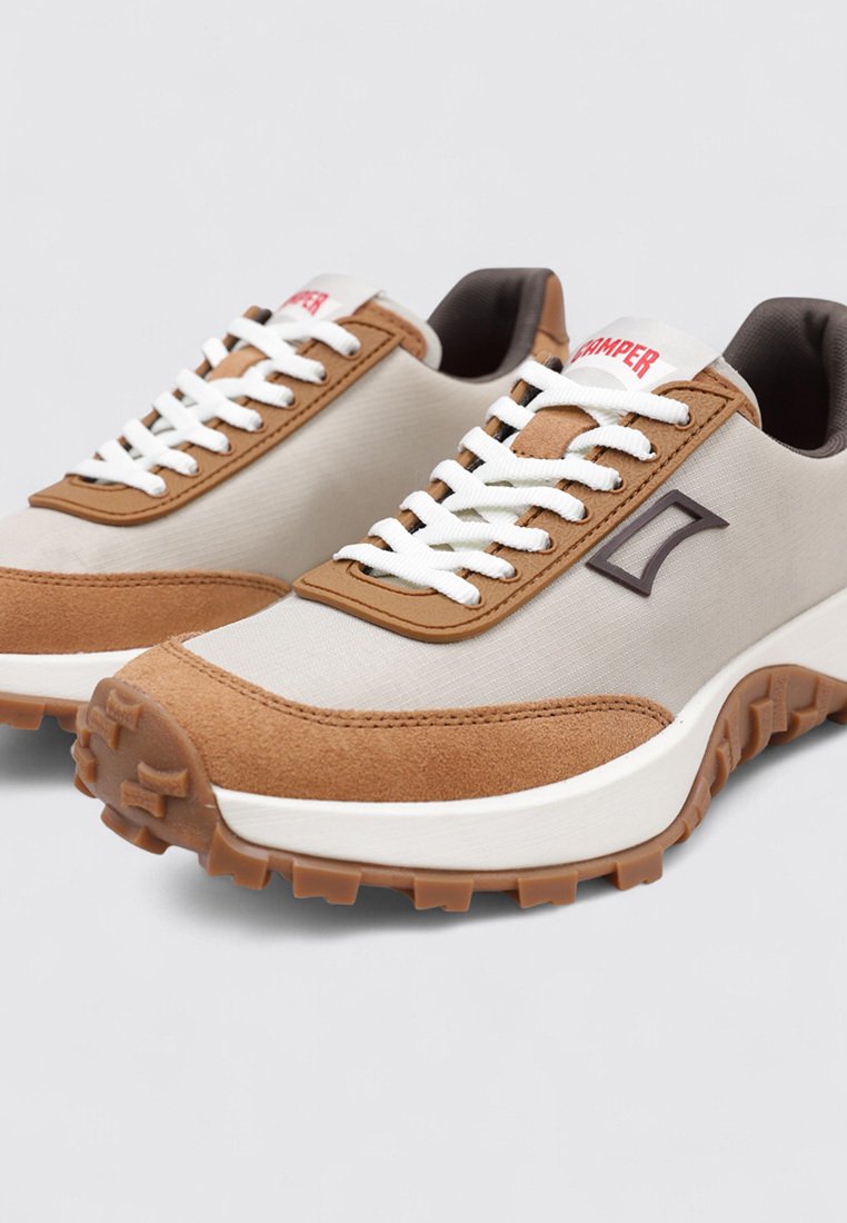 Beige sportieve schoenen met bruin suède accenten, witte veters, rubberen zool en een logo-detail in donkerbruin aan de zijkant.