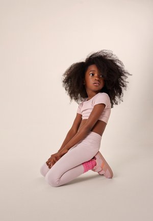 Jeune fille aux cheveux volumineux à genoux, portant un crop top rose pâle, des leggings assortis, des chaussettes roses et des chaussures roses sur un fond uni.
