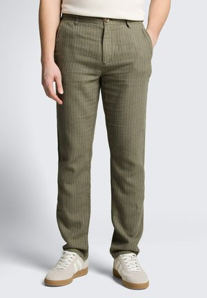Mężczyzna w oliwkowo zielonych spodniach w cienkie paski i beżowych trampkach z białymi paskami, stoi z jedną ręką w kieszeni.