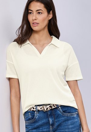 Kobieta ubrana w kremowy, krótki sweter z kołnierzykiem, niebieskie dżinsy oraz pasek w panterkę, stojąca na jednolitym tle.