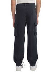 Tumši, brīvi pieguļoši kravas bikses ar elastīgām aprocēm, ar sānu kabatām un gludu auduma tekstūru. Valkātas ar baltām sporta apaviem.