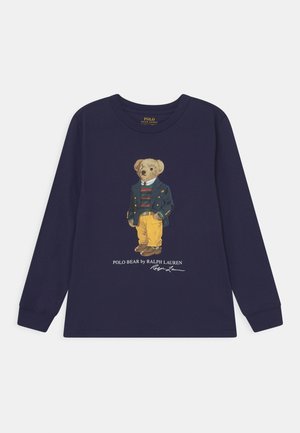 Marineblå langærmet skjorte med Polo Bear-illustration iført blazer, stribet sweater, gule bukser og brune sko, Polo Ralph Lauren-logo nedenunder.