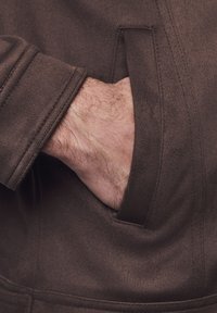 Brązowa, teksturowana kurtka z kieszenią boczną; widoczna ręka w kieszeni, pokazująca delikatne owłosienie i odcień skóry.