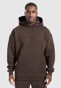 Sudadera con capucha marrón, con bolsillo frontal, corte oversize, y puños y dobladillo acanalados, con el logo en relieve "SMILODOX ORIGINALS". Textura suave.