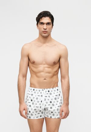 Skjorteløs mand med kort mørkt hår iført hvide badeshorts med sorte abstrakte symboler og logoer, stående foran en ensfarvet baggrund.