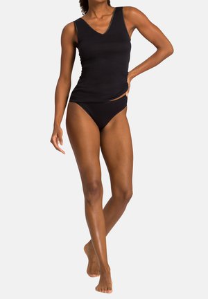 Kvinde står barfodet iført sort ærmeløs top og matchende sort undertøj foran en ensartet lys baggrund.