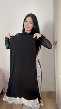 Woman with long black hair holding a black skirt with white lace trim, standing in a minimalist room with light-colored walls and wooden floor.