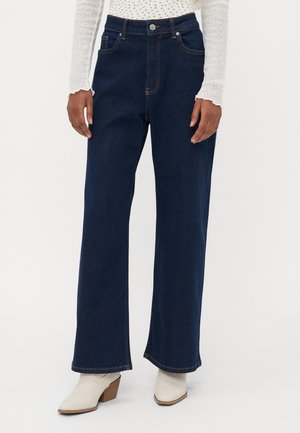 Person iført mørkeblå jeans med vide ben, hvidt strikket langærmet top og hvide ankelstøvler med træhæl.