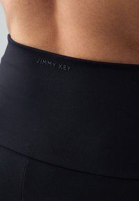 Leggings neri a vita alta con una texture liscia e elastica. Presentano il logo embossato "JIMMY KEY" sulla fascia in vita.