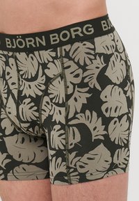 Boxer shorts noirs avec un grand motif de feuilles tropicales beiges, dotés d'une ceinture élastique confortable portant la marque "BJÖRN BORG" en noir.