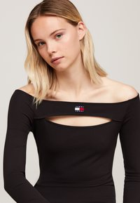 Fekete, hosszú ujjú felső, off-the-shoulder dizájnnal, kivágással a mellen, valamint piros, fehér és kék márkás logóval.