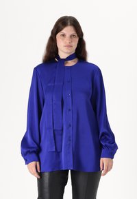 Blå satinbluse med lange ærmer, knapfront og bundet halsdetalje. Glat tekstur, afslappet pasform og knapper ved manchetterne.