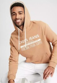 Beżowa bluza z kapturem z białym napisem, kaptur ze sznurkiem, kieszeń z przodu oraz ściągacze u rękawów. Model w białych spodniach, siedzi na białej powierzchni.