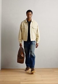 Beige Denimjacke über einem schwarzen Hemd, blauen Jeans und braunen Schuhen. Eine braune Lederreisetasche tragend. Neutrale Hintergrund und Holzfußboden.