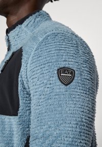 Jachetă din fleece de culoare albastru deschis, cu o suprafață texturată, accente negre, un buzunar cu fermoar și un patch cu logo-ul EA7 pe umărul stâng.
