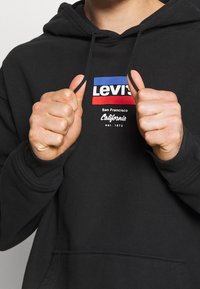 Sweat à capuche noir avec poche kangourou, arborant un logo Levi's en rouge, bleu et blanc. Confectionné dans un tissu doux et texturé.