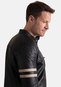 Black leather jacket with a front zip pocket, stand-up collar, and two beige stripes on the left sleeve. Smooth texture and fitted design.