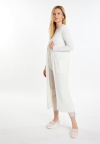 Cardigan lung alb cu textură ribbed, buzunare frontale și deschidere frontală. Purtați peste un top deschis la culoare și pantaloni bej, asortați cu pantofi roz deschis.