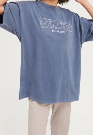 Persoon draagt een oversized blauwe T-shirt met de tekst "ADDICTED TO PARADISE", gecombineerd met lichtbeige broeken tegen een effen achtergrond.