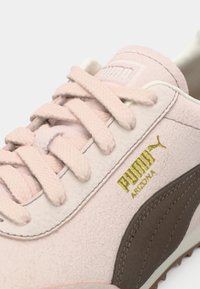 Detailný záber na svetloružovú tenisku Puma Arizona s hrubými šnúrokmi a zlatým logom a nápisom Puma na boku.