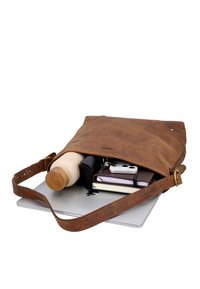 Bolso de hombro de cuero marrón con portátil, botella de agua reutilizable, smartphone, cuadernos y llavero en el interior, parcialmente abierto sobre fondo blanco.