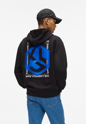 Homme portant un sweat à capuche noir avec un logo bleu et blanc de Karl Lagerfeld Jeans dans le dos, un jean bleu et une casquette en mesh noire, de profil.