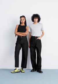 To personer står side om side. Den ene har på seg en svart ermeløs topp og svarte høyhalsede jeans, mens den andre har på seg en grå t-skjorte og baggy svarte jeans.