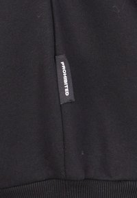 Czarna bluza z tkaniny z ściągaczami na mankietach i dolnym końcu. Posiada naszywkę z napisem "PROHIBITED" na białym tle. Gładka tekstura, casualowy design.